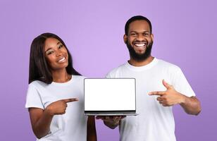 Check This Website. Portrait of happy African American man and woman holding laptop with black blank screen, pointing fingers at computer with empty space for mock up, blue studio background, banner photo