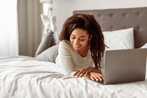 Two Factor Authentication. Portrait of positive black female using phone and laptop in bed, reading message with security code to access financial data online, free space photo