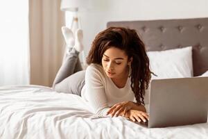 Always In Touch. Portrait of young African American woman lying in bed, using mobile phone and working on laptop computer. Curly black lady reading text message, free copy space photo