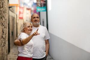 A couple stands together in a narrow street, with one pointing while sharing a joyful moment. Their expressions reflect happiness, surrounded by quaint stone walls and vibrant buildings. photo