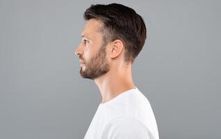 Profile portrait of middle-aged bearded man in white t-shirt over grey studio background, copy space. Side view of handsome confident man posing on gray, standing straight and looking aside photo