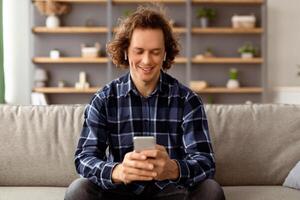 Positive Guy In Earbuds Using Smartphone Listening To Music Online Sitting On Sofa At Home. Man Relaxing Using Mobile Application And Browsing Internet In Living Room Indoor. photo