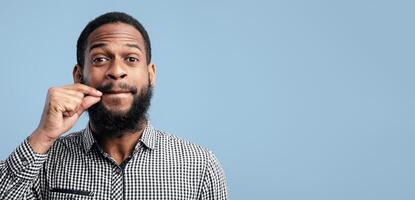 I Won't Say A Word. Black bearded guy making zipping lips gesture with hand, showing he will keep a secret, blue studio photo