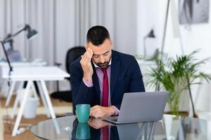 In a modern office, a businessman sits at a glass table, looking stressed while resting his forehead on his hand. A laptop is in front of him, and he holds a coffee mug in his other hand. photo