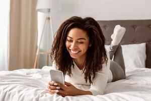 Mobile Phone User. Portrait of happy African American woman lying in bed and smiling while reading text message from someone, holding and using smartphone, chatting with lover or browsing social media photo