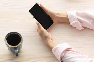 Above top high angle view of lady holding and using smart phone with black empty screen for mock up design, having breskfast, drinking coffee in the morning, searching for recipes online, free space photo