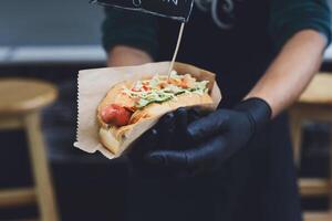 Fresh hot dog with grilled sausage, served outdoors in craft paper. Cookout american bbq food. Hot-dog with meat and vegetables salad filling closeup in chef's hands. Street food, fast food. photo