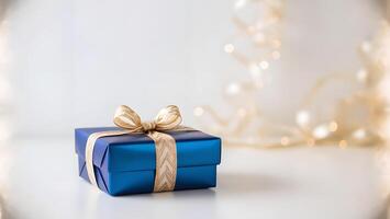 Blue gift box with gold ribbon and bow on white surface. Blurred festive background with soft lights photo