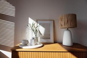 A wooden chest of drawers and a window with louvered shutters. An elegant table lamp, poster frame mock-up, a tray with a glass jug and a vase, scented candles and cup. photo