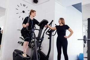 Elderly plump woman in electrical muscle stimulation suit is training on elliptical trainer with her personal coach in gym or fitness club. photo