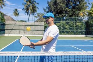 A young man in a cap and sunglasses throws a yellow ball and holds a racket. An athlete plays pickleball outdoors. photo