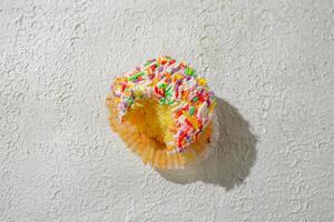A colorful cupcake bitten topped with vanilla frosting and a generous amount of rainbow sprinkles, set against a textured white background for a clean and appealing look photo