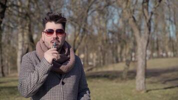 A young handsome bearded hipster man vaping an electronic cigarette in the park in the spring. Close up. video