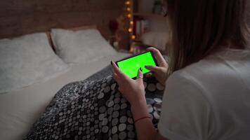 Young woman relaxing on her bed with her mobile phone. Mockup of girl using Horizontal green screen of smartphone. Female clicks finger on touchscreen of cellphone. video