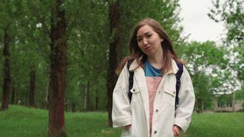 Beautiful woman smiles walking along path surrounded by foliage and trees. Front view of female with backpack walking at the summer park. video