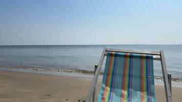 Beachside Serenity A striped beach chair reclines on a sandy shore, facing a calm sea. The blue sky and tranquil water create a serene atmosphere, inviting relaxation and escape. video