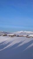 Snow blankets the charming wooden cabins nestled among tall evergreen trees. Majestic mountains rise in the background under a bright blue sky, capturing the tranquility of winter. Vertical footage video