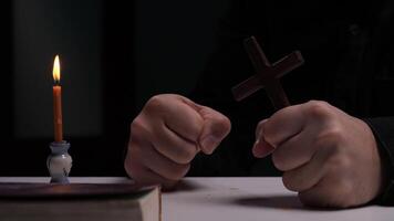 Priest holding cross and clenching fists on table with candle and bible video
