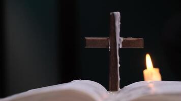 Wooden cross covered with candle wax standing on open book with lit candle in background video