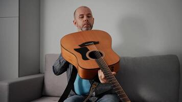 A middle-aged man with a short beard and shaved head is sitting on a gray couch, holding an acoustic guitar. The man is examining the guitar closely, possibly tuning it or inspecting its build. video