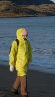 Tourist, wearing raincoat, walks along coast, enjoying waves of ocean and view of northern landscapes typical of Scandinavian countries. video