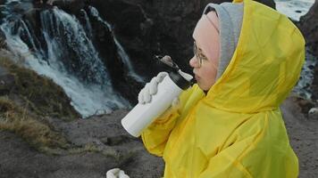 Young woman, dressed in warm, waterproof raincoat, stands against backdrop of beautiful waterfall. She is drinking hot tea from thermos, trying to stay warm in cold northern climate of tundra. Portrait. video