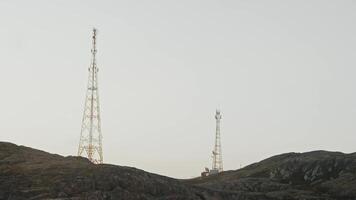 Two tall telecommunication towers are located on an elevated area of rocks in middle of cold northern tundra. They transmit radio signals of different frequencies. video