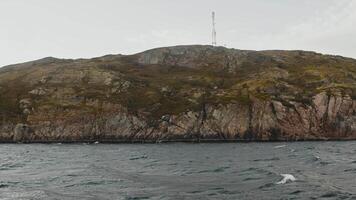 A view of a steep, rocky shore covered in sparse grass and moss, washed by sea waves, in a cold northern landscape. video