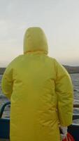Back of fisherman wearing bright yellow waterproof raincoat, with hood, sailing on fishing boat in midst of cold and windy weather. video