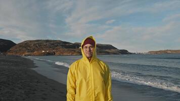 Young fisherman in colorful warm waterproof raincoat stands on cold sandy beach against backdrop of northern sea bay and rocky hills video