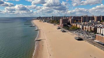 Aerial view of urban beachfront with buildings and blue ocean - A stunning aerial view of a sandy urban beachfront with calm blue ocean waves, buildings along the coast, and a clear sky dotted with video