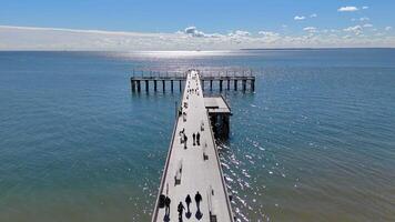 Sunny Day on a Scenic Ocean Pier. Aerial view of a pier extending into the sparkling ocean under a bright blue sky, with people strolling and relaxing. video
