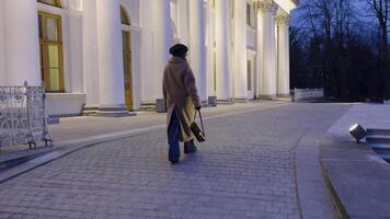 en kvinna i en beige täcka promenader på en kullersten väg mot en ljust upplyst historisk herrgård. de elegant arkitektur och skymning miljö skapa en lugn och tidlös atmosfär video