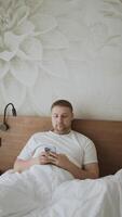 Happy Caucasian young man using smartphone for calls, social networks, mobile applications online in a bed at hotel video