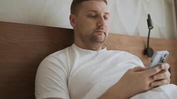 Happy Caucasian young man using smartphone for calls, social networks, mobile applications online in a bed at hotel video