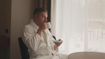 Young caucasian man in a white coat drink coffee look at the snow view throw the window. video