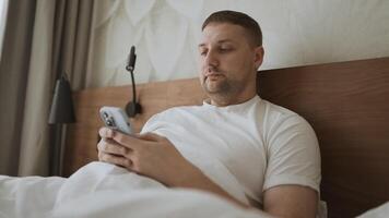 Happy Caucasian young man using smartphone for calls, social networks, mobile applications online in a bed at hotel video
