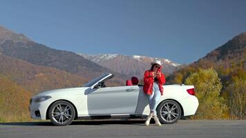 Smiling woman is browsing in phone standing near convertible car in mountains. video