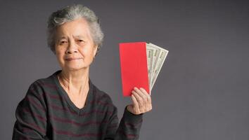 An elderly Asian woman in a sweater holding a red envelope with a US dollars banknote and looking at the camera with a smile while standing on gray background photo