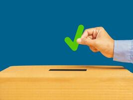 Hand holding a green check mark symbol overhead the voting box at place election with a blue background photo