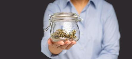 Close-up of a hand holding a glass jar with dry cannabis buds while standing on a gray background photo