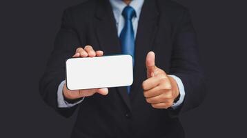 A businessman holding a horizontal mobile phone with a blank white screen and showing thumbs up while standing on a gray background photo