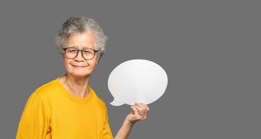 Senior woman in an orange casual holding a blank speech bubble and looking at the camera with a smile while standing on a gray background photo
