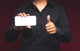A businessman holding a horizontal mobile phone with a blank white screen and showing thumbs up while standing on a red background photo