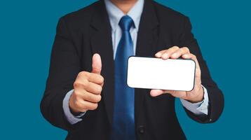 A businessman holding a horizontal mobile phone with a blank white screen and showing thumbs up while standing on a blue background photo