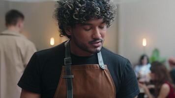 Arabian Indian Latino man chef waiter in apron serving customer in cafe friendly smiling male guy talk with client in restaurant serve food order writing at digital tablet modern service coffee shop video