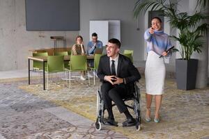 In a professional office setting, a businesswoman helping her director, who is sitting in a wheelchair and using a tablet, while their colleagues collaborate in the background. photo