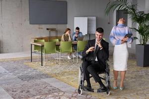 In a professional office setting, a businesswoman helping her director, who is sitting in a wheelchair and using a tablet, while their colleagues collaborate in the background. photo