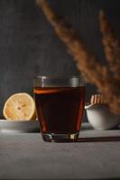 Glass with black tea and slice of lemon on the table, honey spoon and lemon in the background. Vertical photo. Breakfast and morning hot beverage concept. Dark mood photo