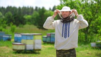 Portrait of beekeeper on apiary. Apiarist wearing protective hat. Man in embroidered shirt puts on hat to protect from bees and smiles on camera. video